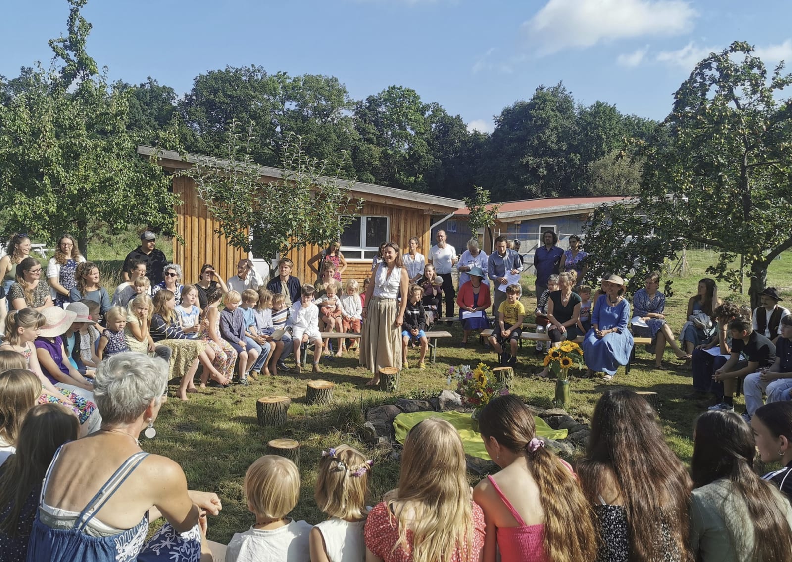 Viele Menschen sitzen und stehen im Halbkreis vor einem Holzhaus unter Obstbäumen